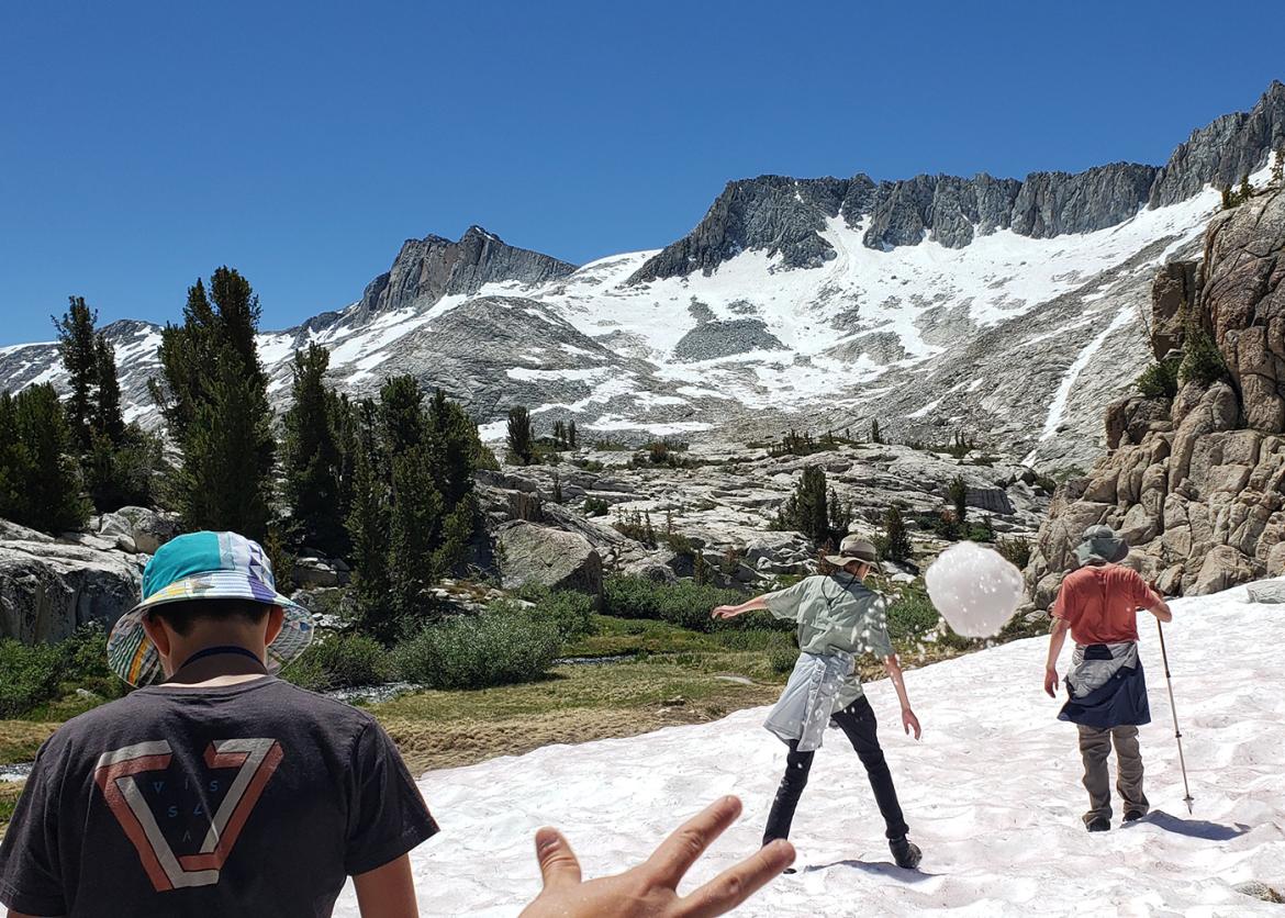 Teen boy throws snowball at fellow participant with beautiful mountain scenery in the background.