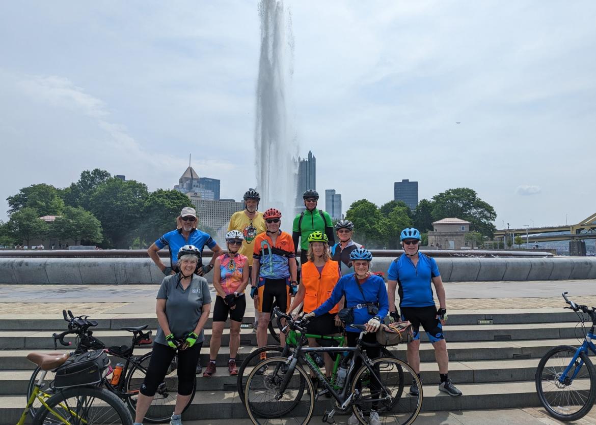 A group of colorful bikers smiling with their bikes and a fountain in the background
