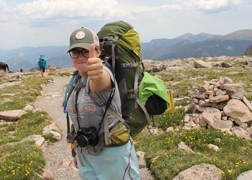 Teen girl wearing backpacking gear gives thumbs-up to camera on a rocky trail.