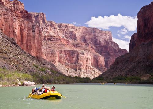A crowded inflatable raft rows across the water in front of a canyon.
