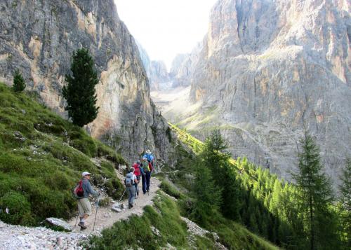 Five hikers walk on a trail on a steep grassy slope, overlooking sheer rocky mountainsides.
