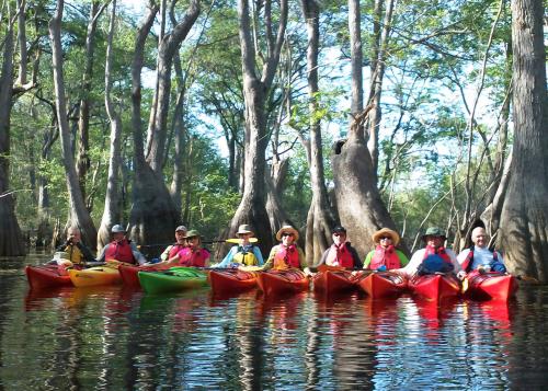 Ten smiling people in kayaks float in a line, holding onto each other's boats to keep in place.