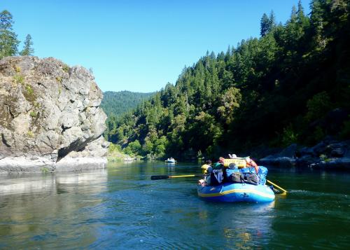 Two inflatable rafts loaded with supplies paddle down a river bordered by forest on one side and rocks on the other.