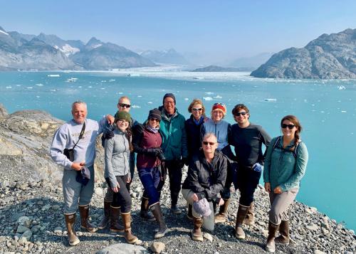 A group of ten smiling people stand in front of a view of mountains and icy waters.