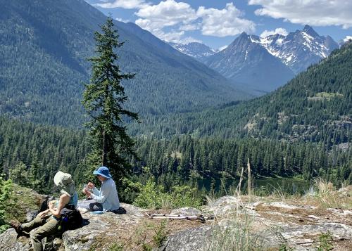 Women pause for a rest along the hiking trail, overlooking tree-covered hills and a snow-capped mountain