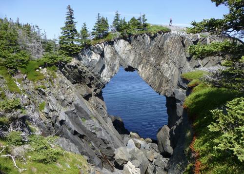 Woman walking over natural rock archway, stretching over the ocean on Newfoundland coast